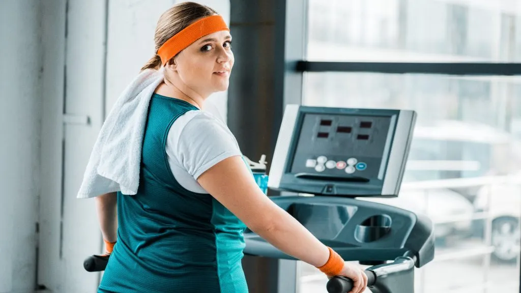 exhausted overweight girl with towel on shoulders looking at camera on treadmill in gym