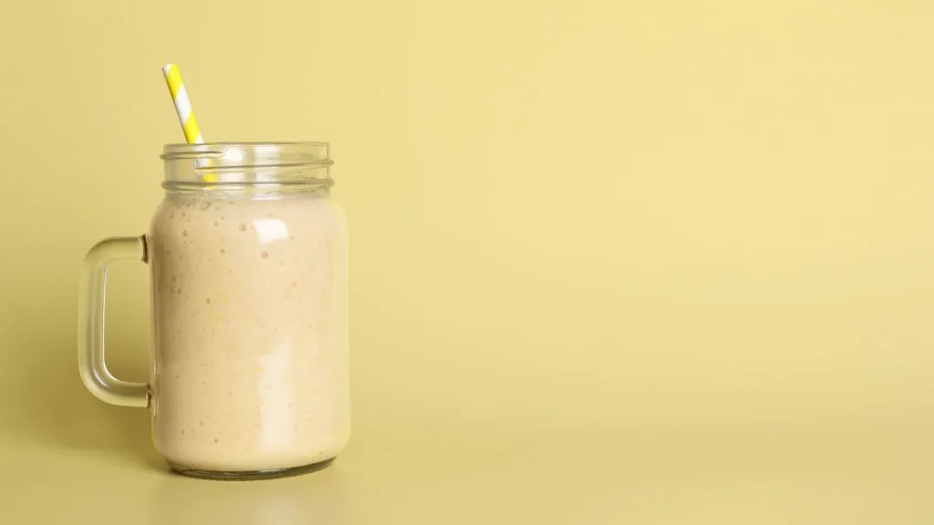 Mason jar of tasty peanut butter smoothie with straw on a beige background.
