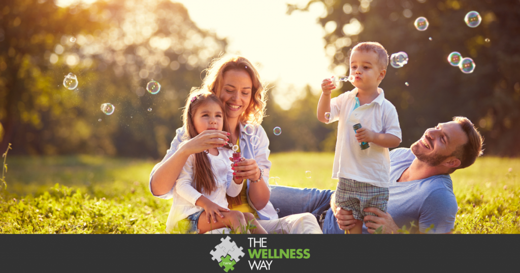 a family of parents and two children sitting in a field of grass and blowing bubbles together.