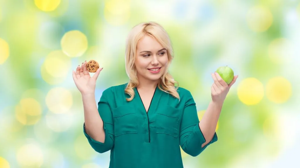 Young woman holding a cookie and green apple. Looking at the apple.