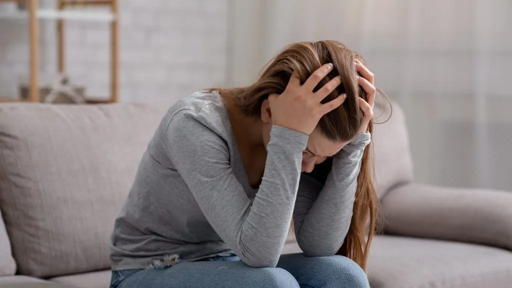 Unhappy young woman with head in her hands sitting on couch, crying and feeling depressed.