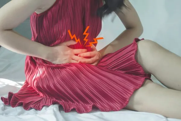 Asian girl wearing a pink dress has severe abdominal pain and is sitting on a bed.
