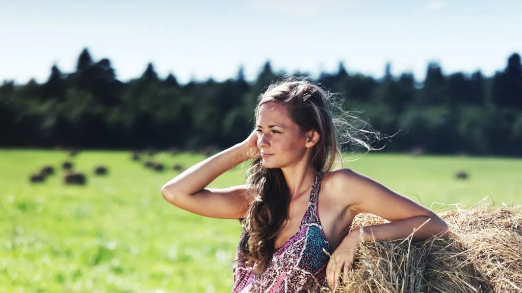 Portrait of a beautiful young woman sitting on a haystack in a field with cows.