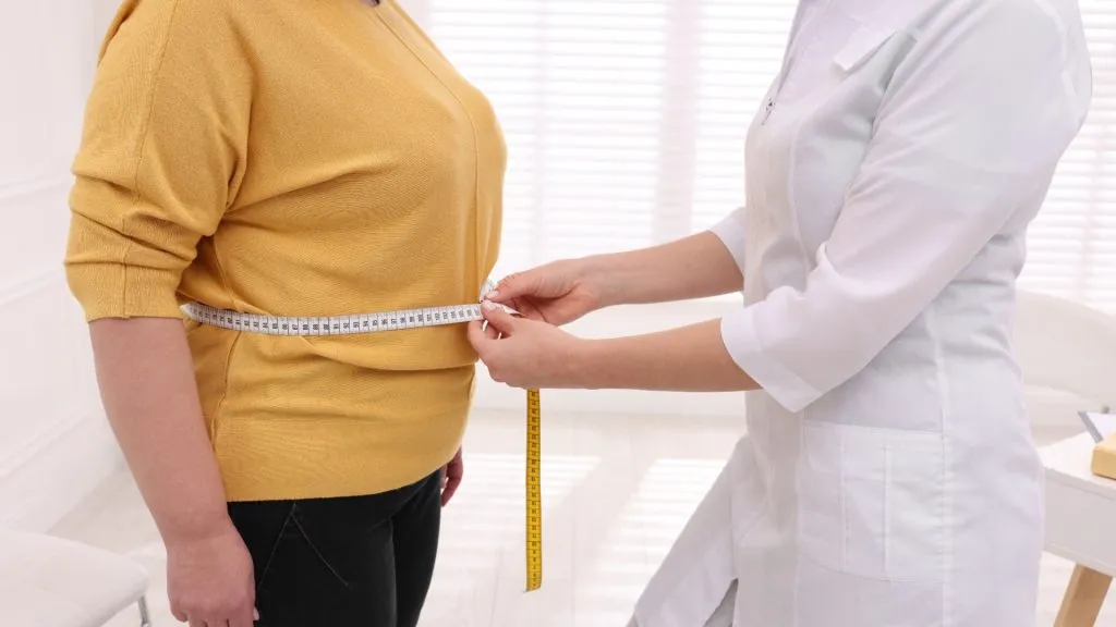 Doctor measuring waist of overweight woman in clinic, closeup. Weight loss concept
