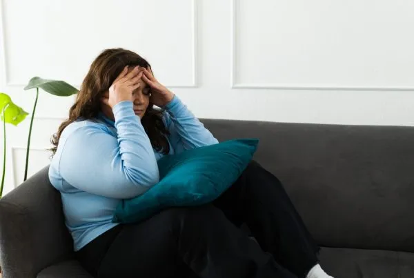 Overweight young woman sitting on sofa and covering face with pillow at home.
