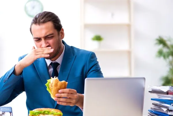 Young male employee having breakfast at workplace stressed and stomach pain