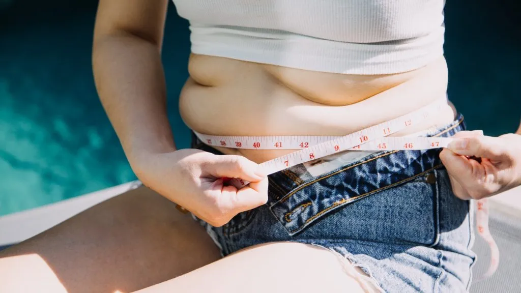 Young woman holding excess belly fat with a measuring tape.